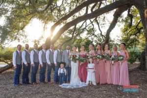 Outdoor luxury wedding ceremony by the water featuring a large wedding party wearing designer bridesmaid dresses and formal cowboy attire