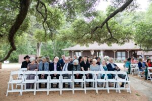 Wedding guests are seated at an outdoor wedding venue under the shade of a historic oak tree with views of the lake